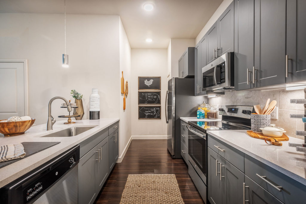 Long shot of galley kitchen with large single basin sink with gooseneck faucet, grey cabinets, stainless steel appliances and hardwood floors.