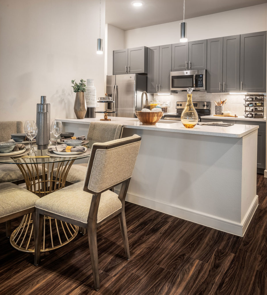 Kitchen interior with stainless steel appliances, marble counters, tile backsplash, gray cabinets, pendant lighting and glass table with seating for 4.