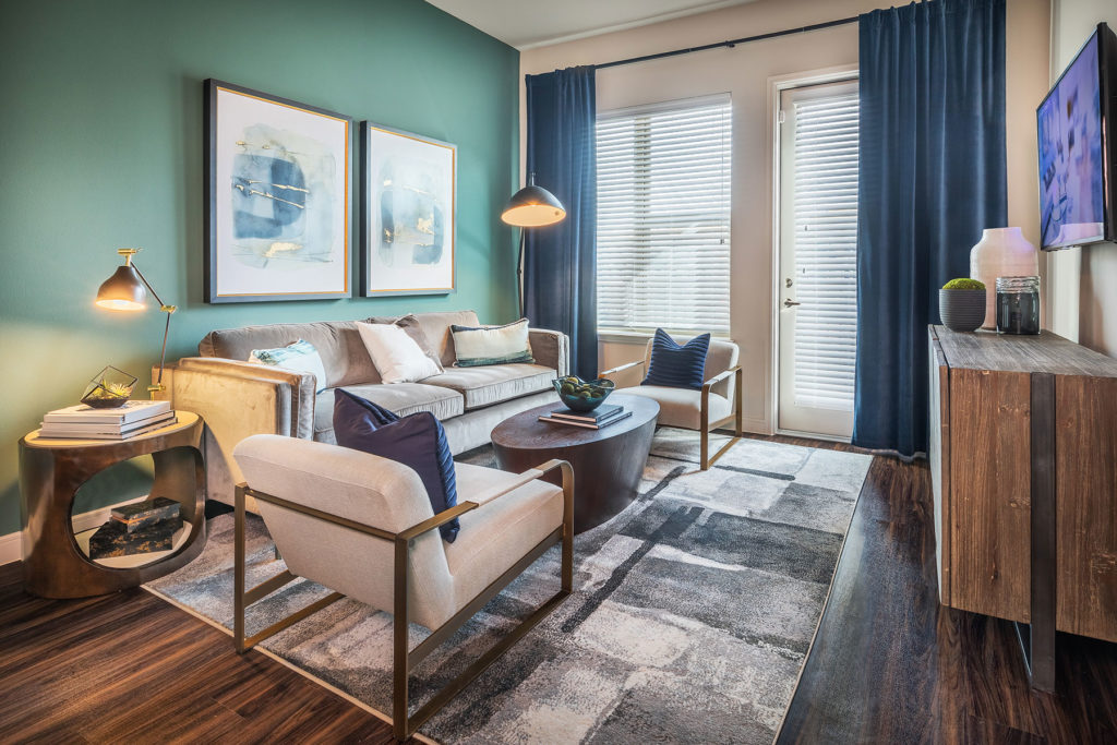 Living room interior with large window and door leading to patio, couch and chair seating with coffee table and cut out side tables, teal accent wall, wooden floors, wall mounted TV with wooden entertainment stand and abstract watercolor artwork.
