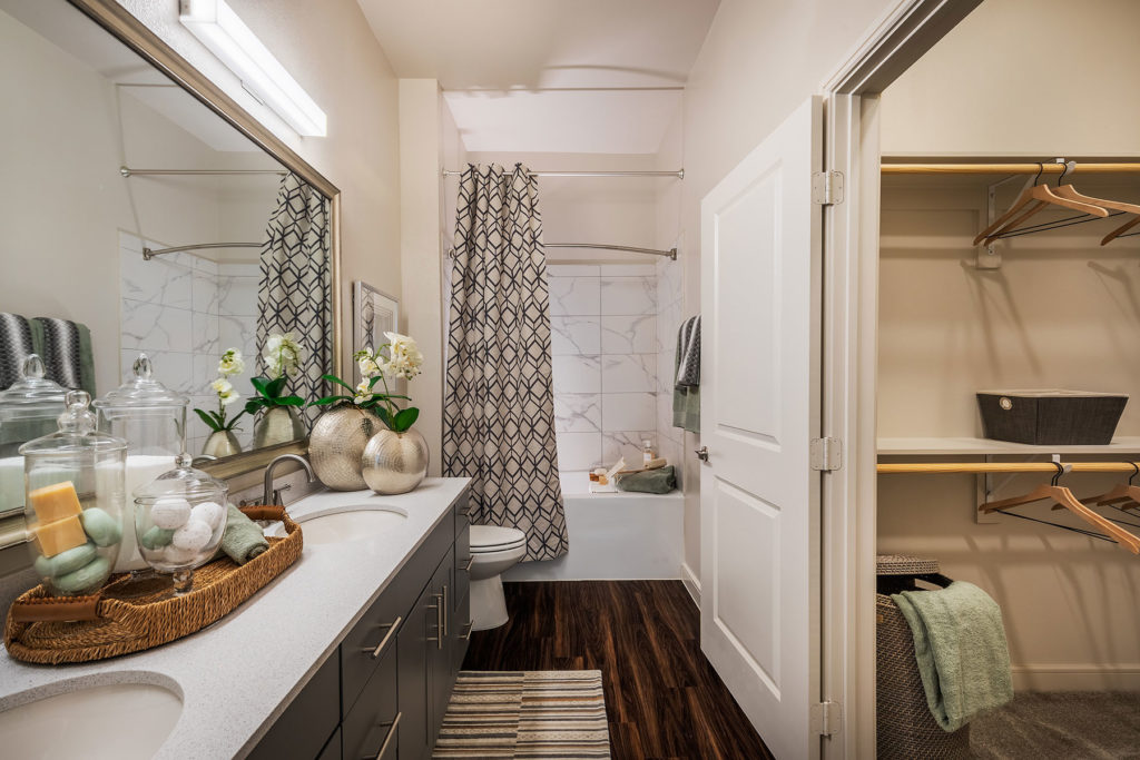 framed mirror, dual sink vanity, wood floors and shower tub combo on the left with walk in closet on the right.