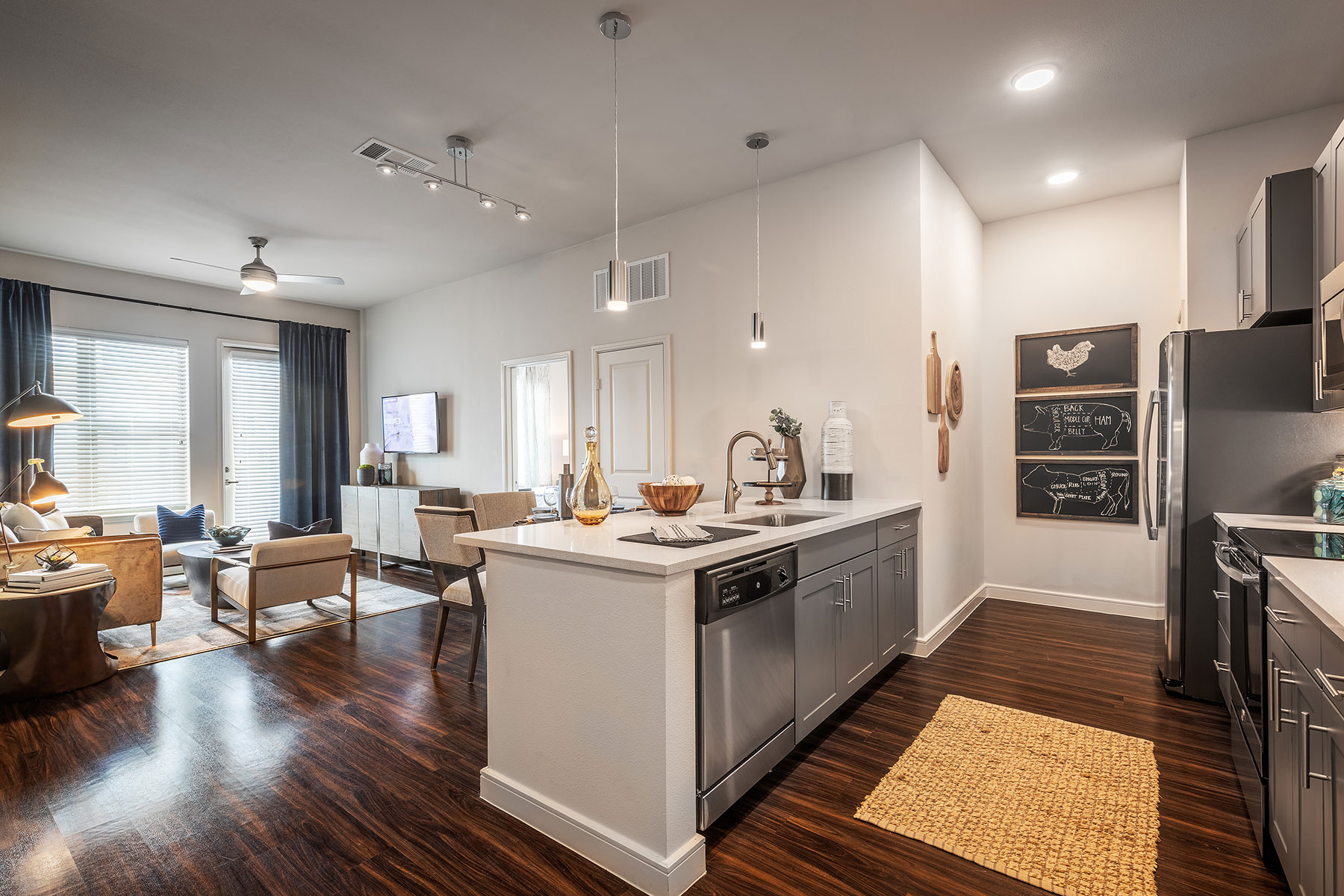 Wide shot of the living area with large windows and comfortable seating, beautiful wooden floor, and kitchen with stainless steel appliances, pendant lights, and farm animal artwork.