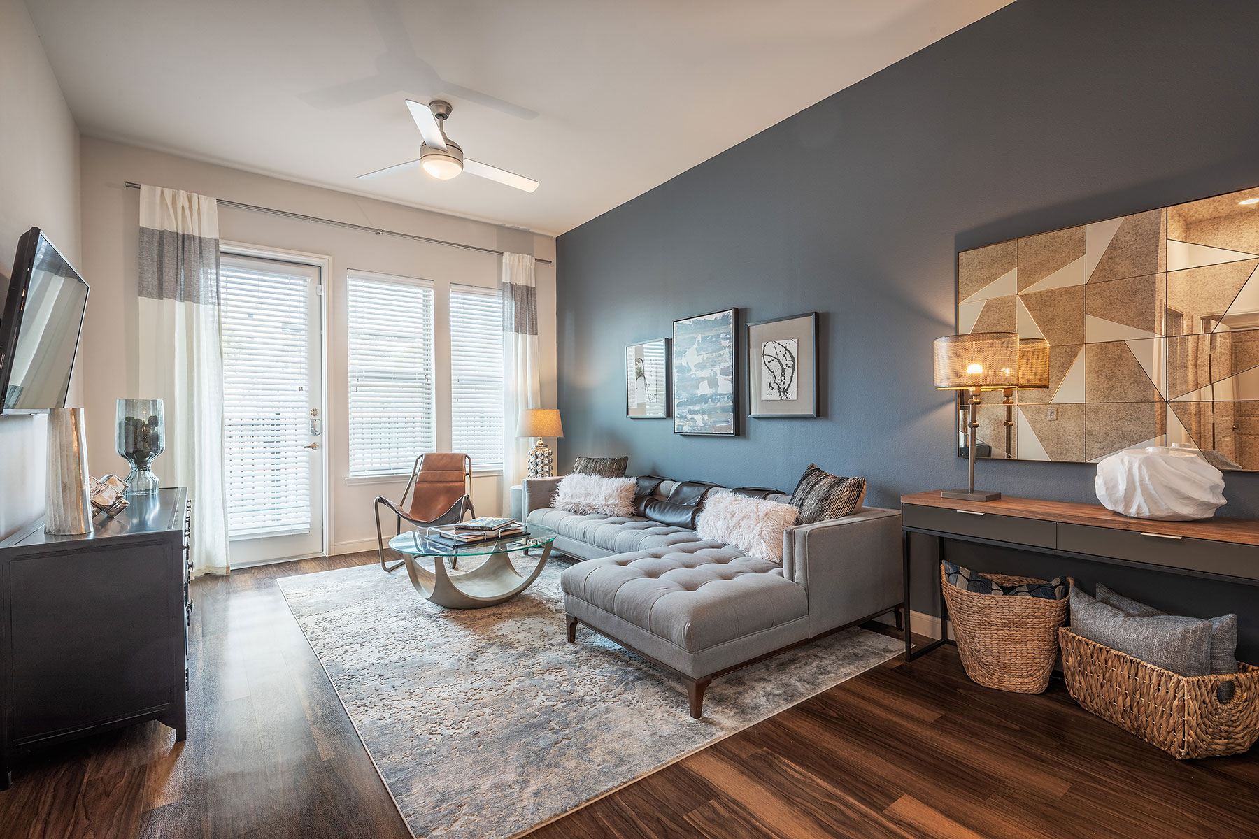 Living room with L shaped sectional couch, glass round coffee table, wall mounted TV, windows and patio door, gray accent wall, fuzzy pillows, long side table with wicker baskets underneath and contemporary artwork hanging on the wall.