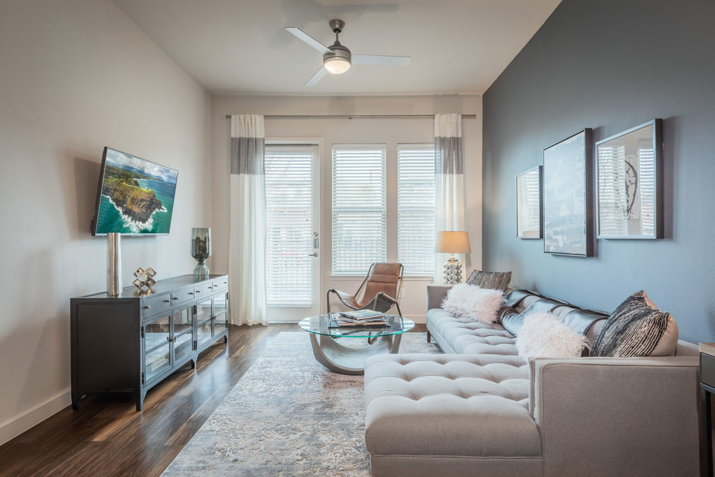 Living room with L shaped sectional couch, glass round coffee table, wall mounted TV, windows and patio door, gray accent wall and fuzzy pillows.