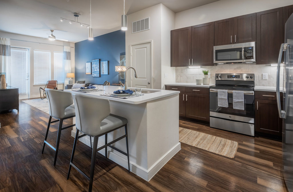 Kitchen interior with island, bar seating, wooden cabinets, stainless steel appliances, and view of the living area in the back