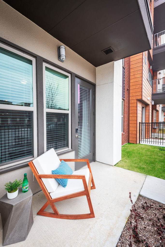 Small patio area with modern wooden rocking chair with plush cushions with small geometric side table and gray door leading inside.
