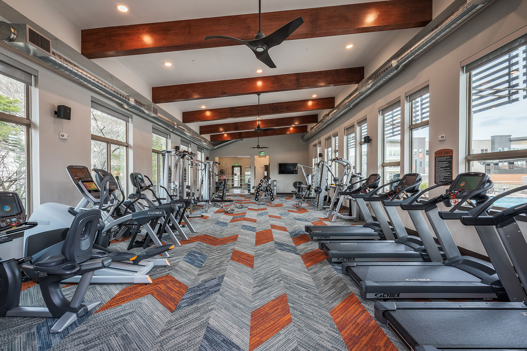 Wide shot of the fitness center with a variety of cardio machines, free weights, wall mounted TV and windows outlooking to the pool.