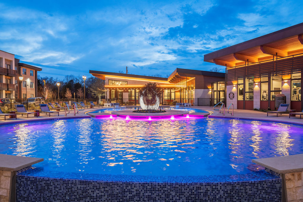 Round view of the pool at night with blue and pink lights with view of the clubhouse in the background.