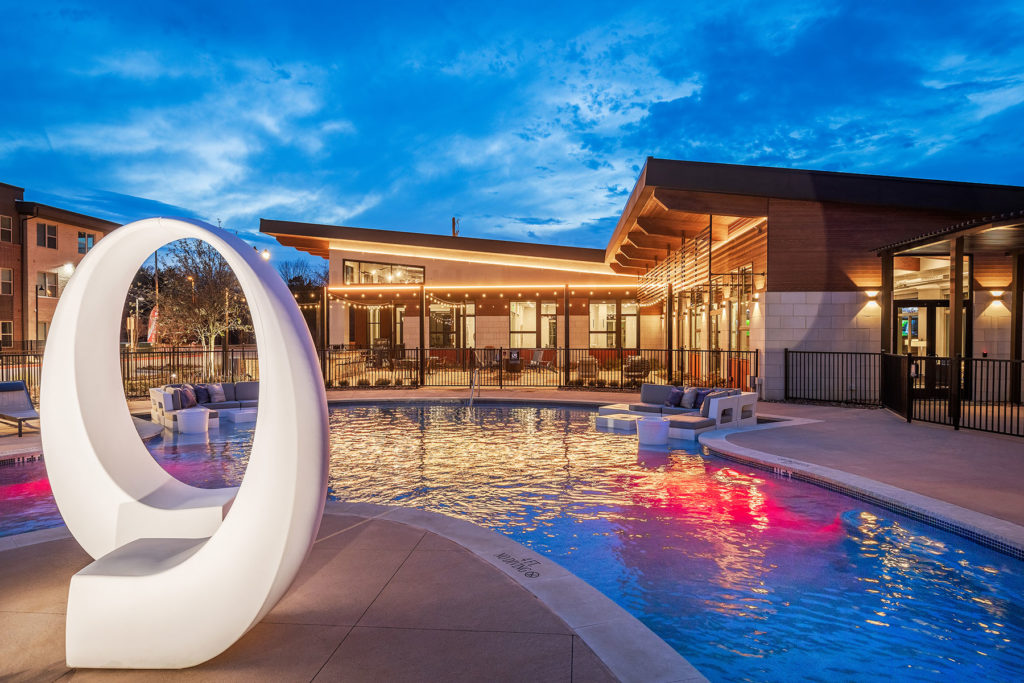Night time pool photo with close up of white oval sculpture, colorful pool lights, patio seating and clubhouse in the background.