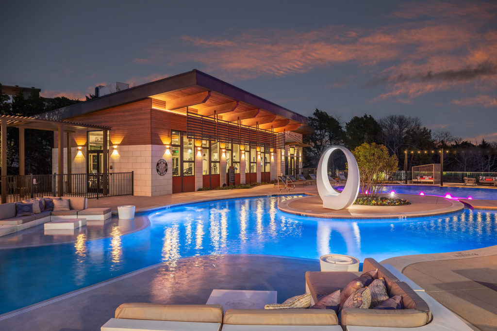Night time pool photo with close up of white oval sculpture, colorful pool lights, patio seating and fitness center in the background.