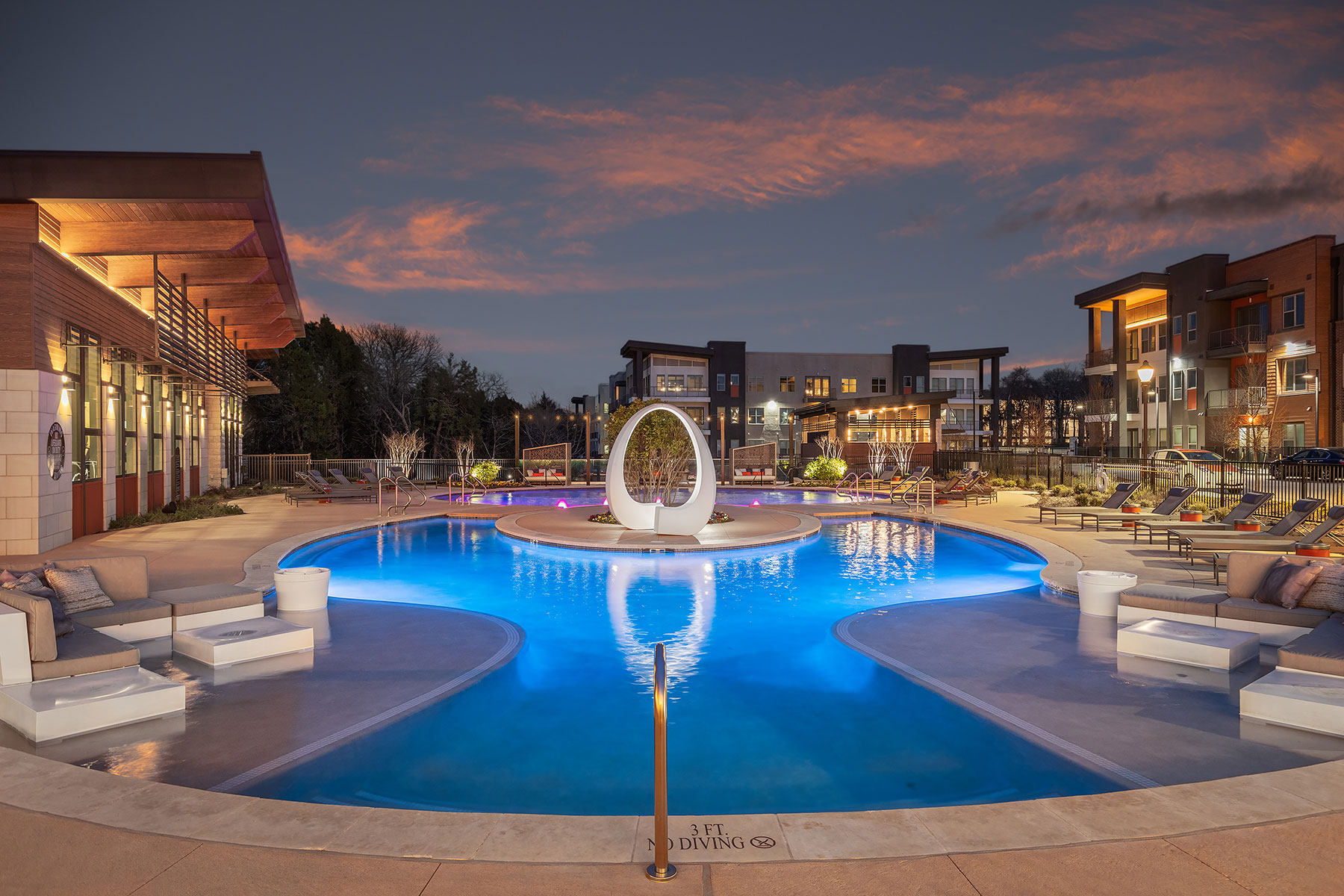 Night time pool shot with oval architecture sculpture, lounge seating, and lighted buildings in the background.