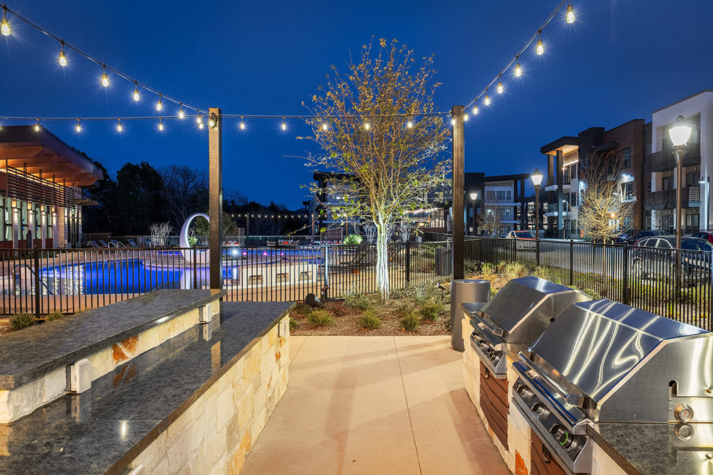 Stainless steel gas grills and outdoor kitchen prep area with string lights by the pool.