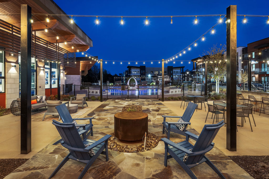 Outdoor pool patio seating area with firepit, string lights and view of the pool at night.
